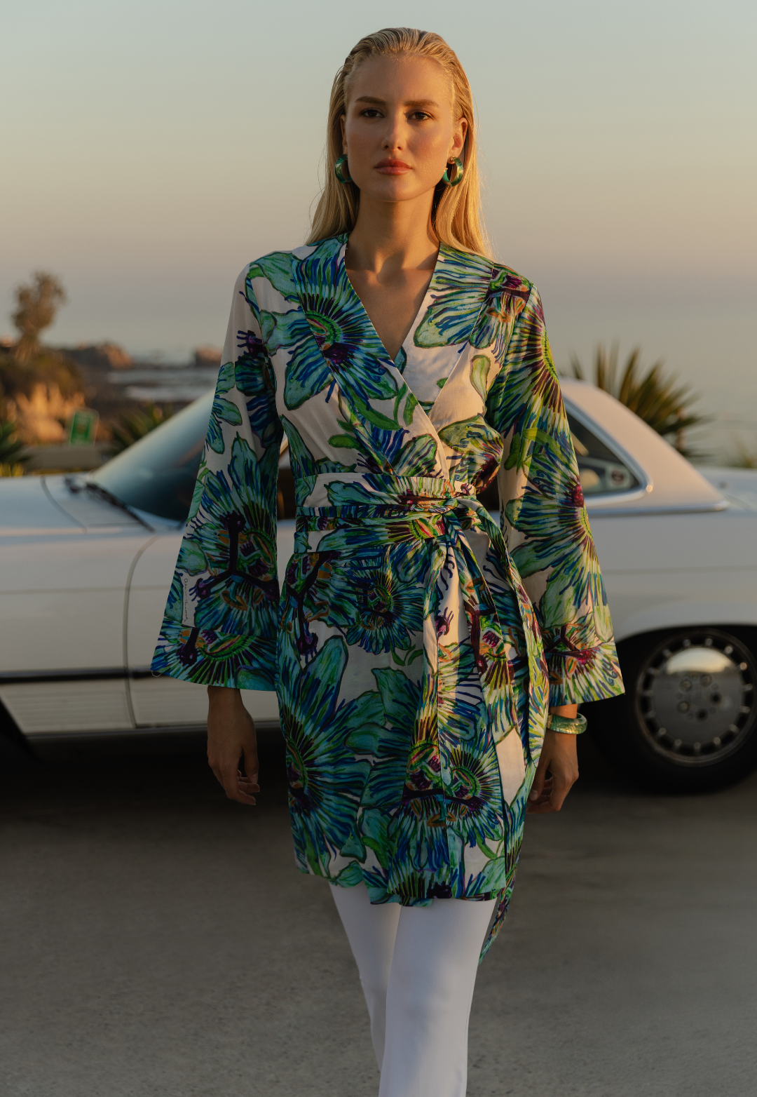 Woman wearing a colorful floral kimono and white pants standing in front of a white car.