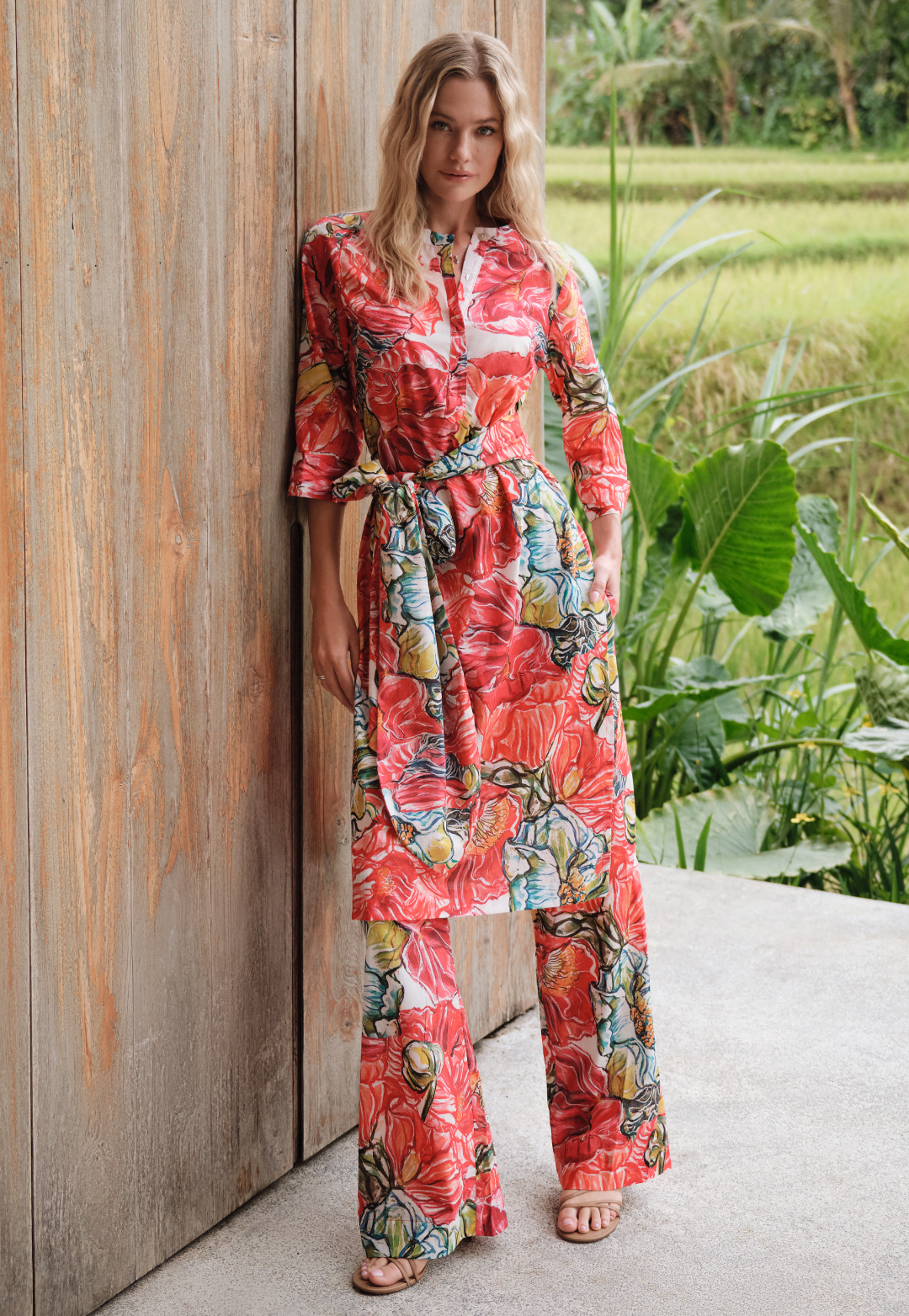 Woman in a colorful floral tunic and pants standing against a wooden wall with greenery in the background