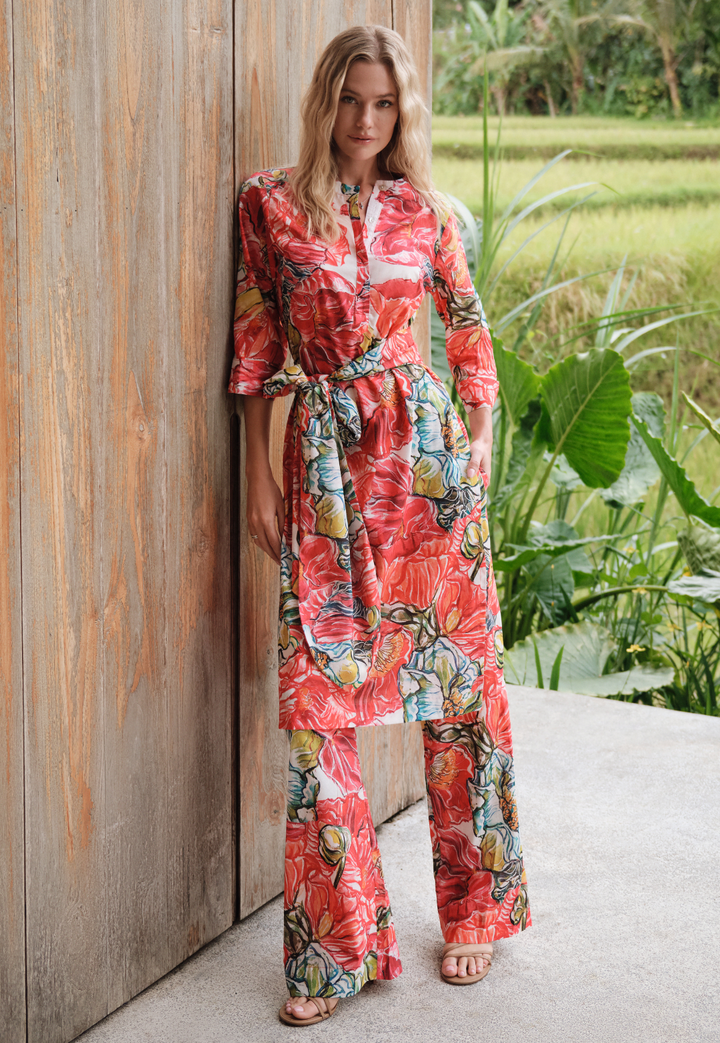 Woman in a colorful floral tunic and pants standing against a wooden wall with greenery in the background