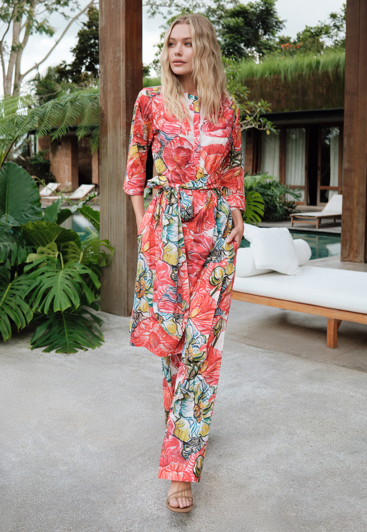 Woman in a colorful floral pants and tunic walking outdoors with greenery and a building in the background