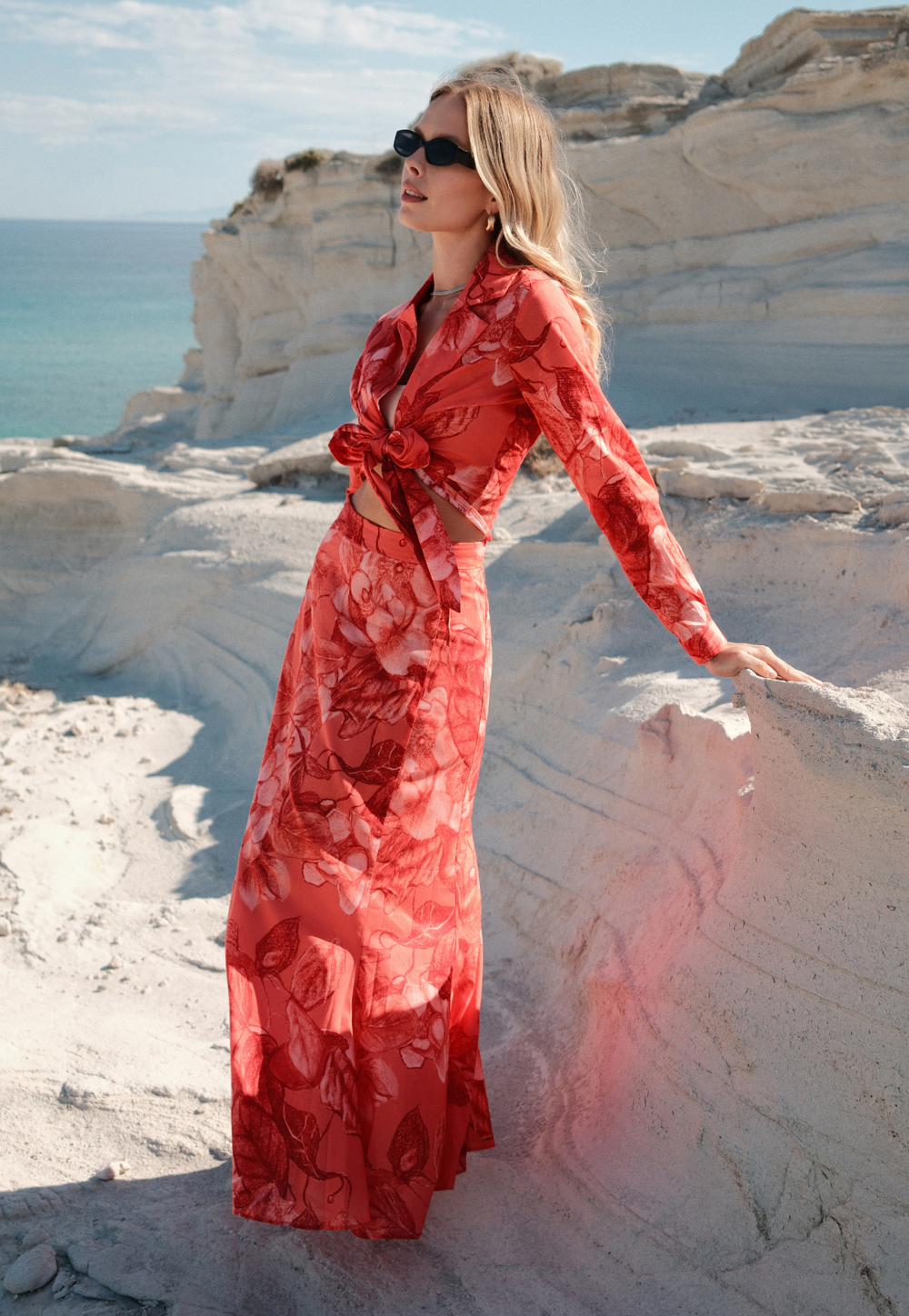 Woman in a red floral dress standing on a rocky coastal landscape.