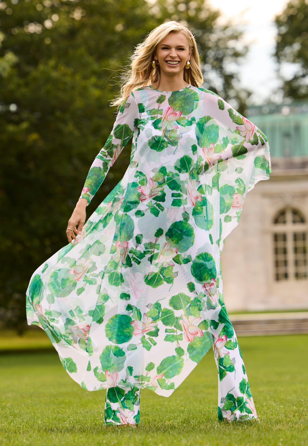 Woman wearing a floral mesh kaftan standing outdoors with greenery and a building in the background