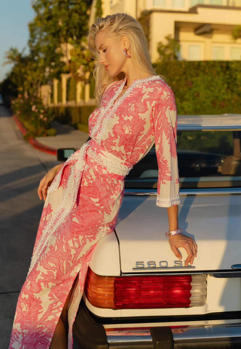 Woman in a pink and white patterned dress leaning against a vintage car.