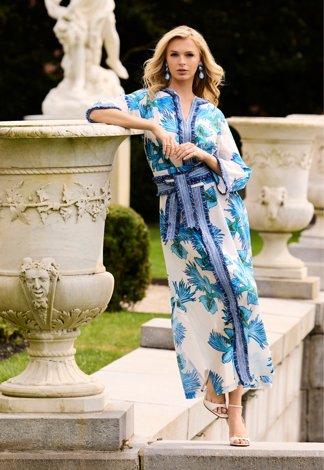 Woman in a blue and white floral dress standing in an elegant outdoor setting with classical sculptures.