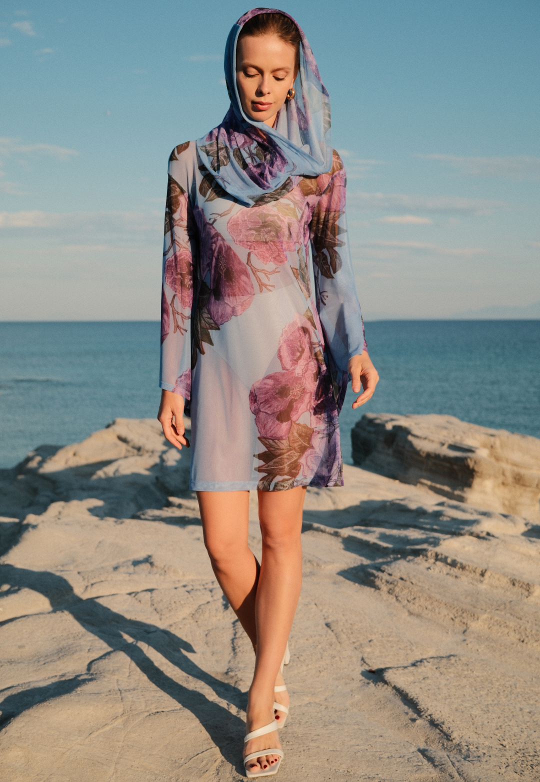 Woman in a floral dress and hood walking on rocks by the ocean.