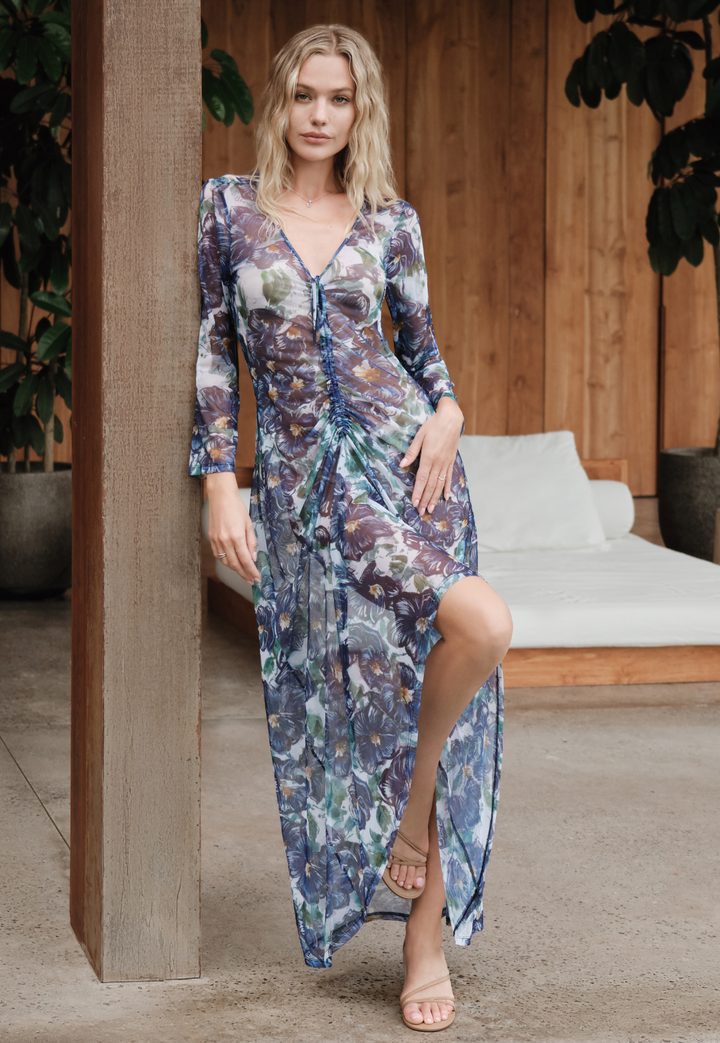 Woman wearing a floral dress standing in an outdoor setting with plants and a wooden structure.