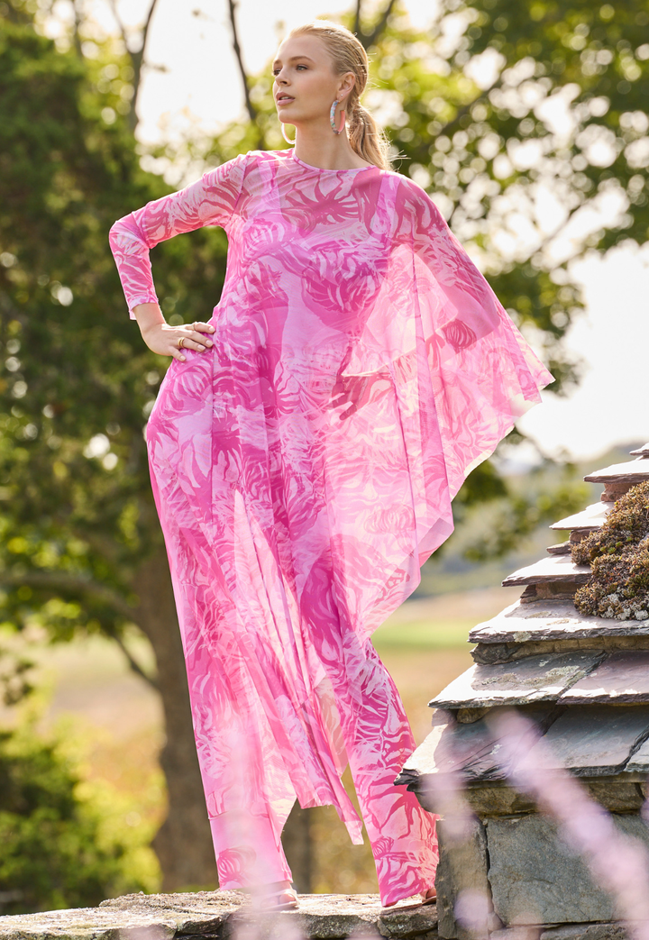 Woman wearing a pink floral mesh poncho over matching tank top and pants standing outdoors with greenery in the background