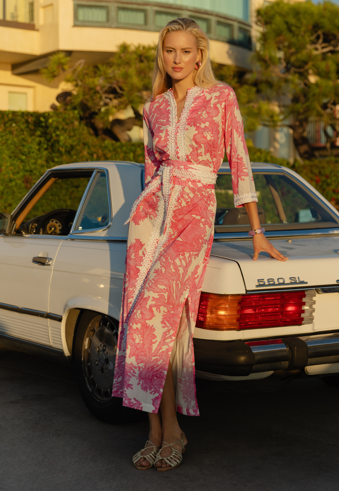 Woman in a pink and white patterned dress standing next to a vintage car.