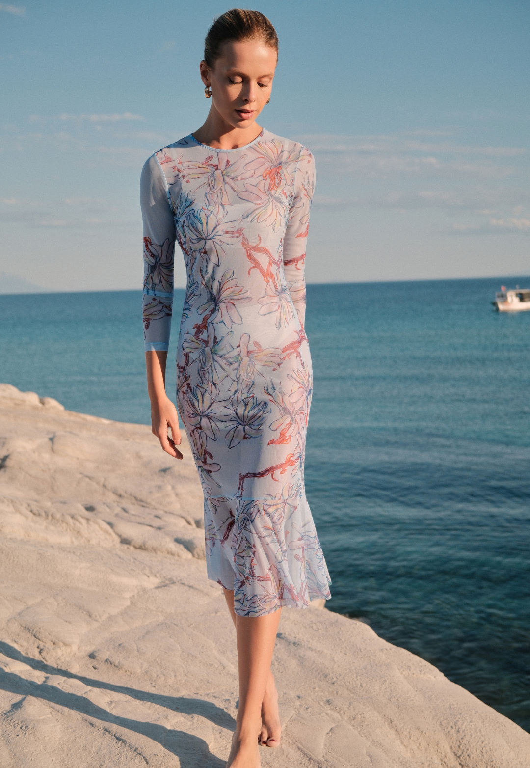 Woman in a floral dress standing on a rocky shore with ocean and sky in the background