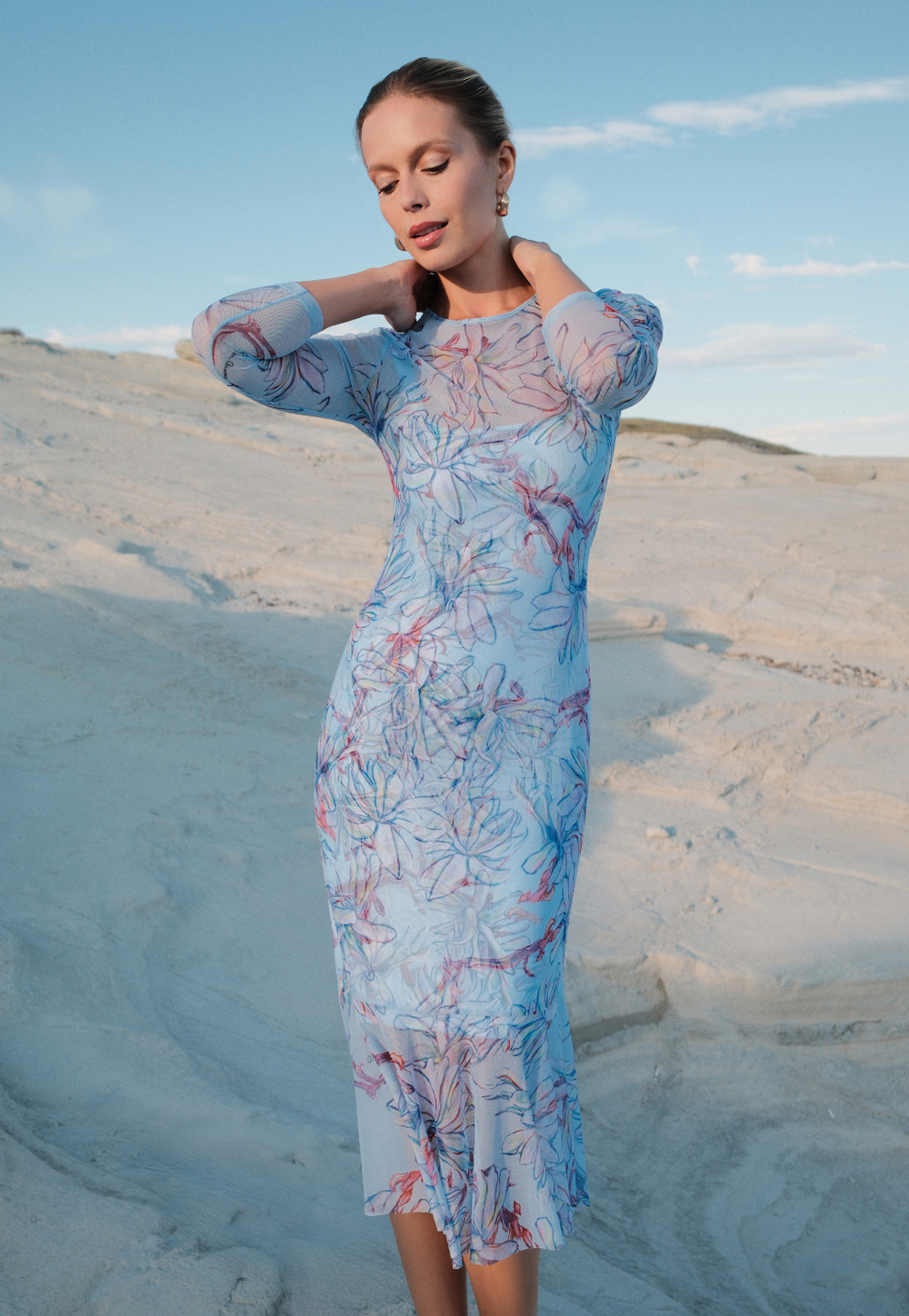 Woman in a floral dress standing on a sandy landscape with blue sky.