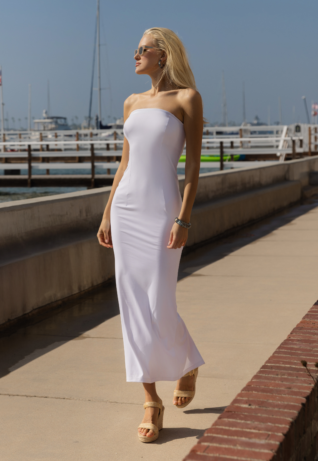 Woman in a white strapless dress standing on a dock with boats in the background.