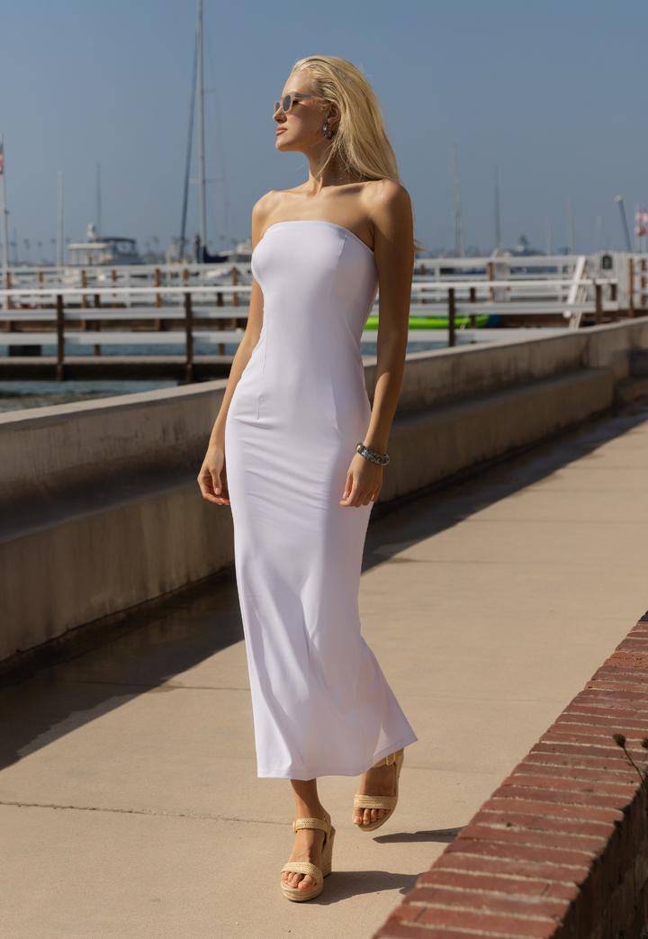 Woman in a white strapless dress standing on a dock with boats in the background.