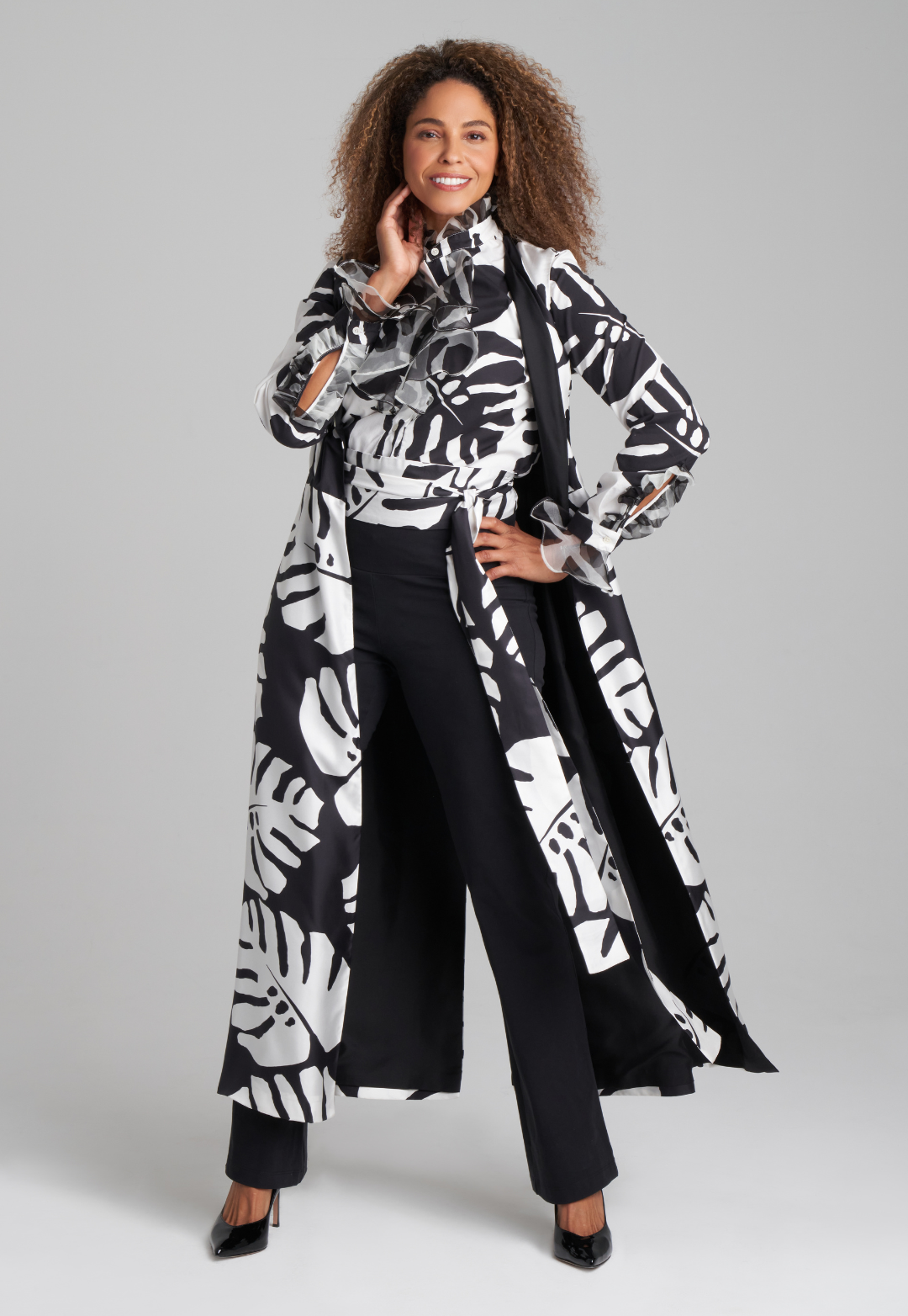 Woman wearing cotton turtleneck black and white palm leaf printed blouse with ruffle sleeve and collar details with italian stretch cotton pant in black with black and white printed obi belt sash with reversible long vest by Ala von Auersperg for holiday 2022