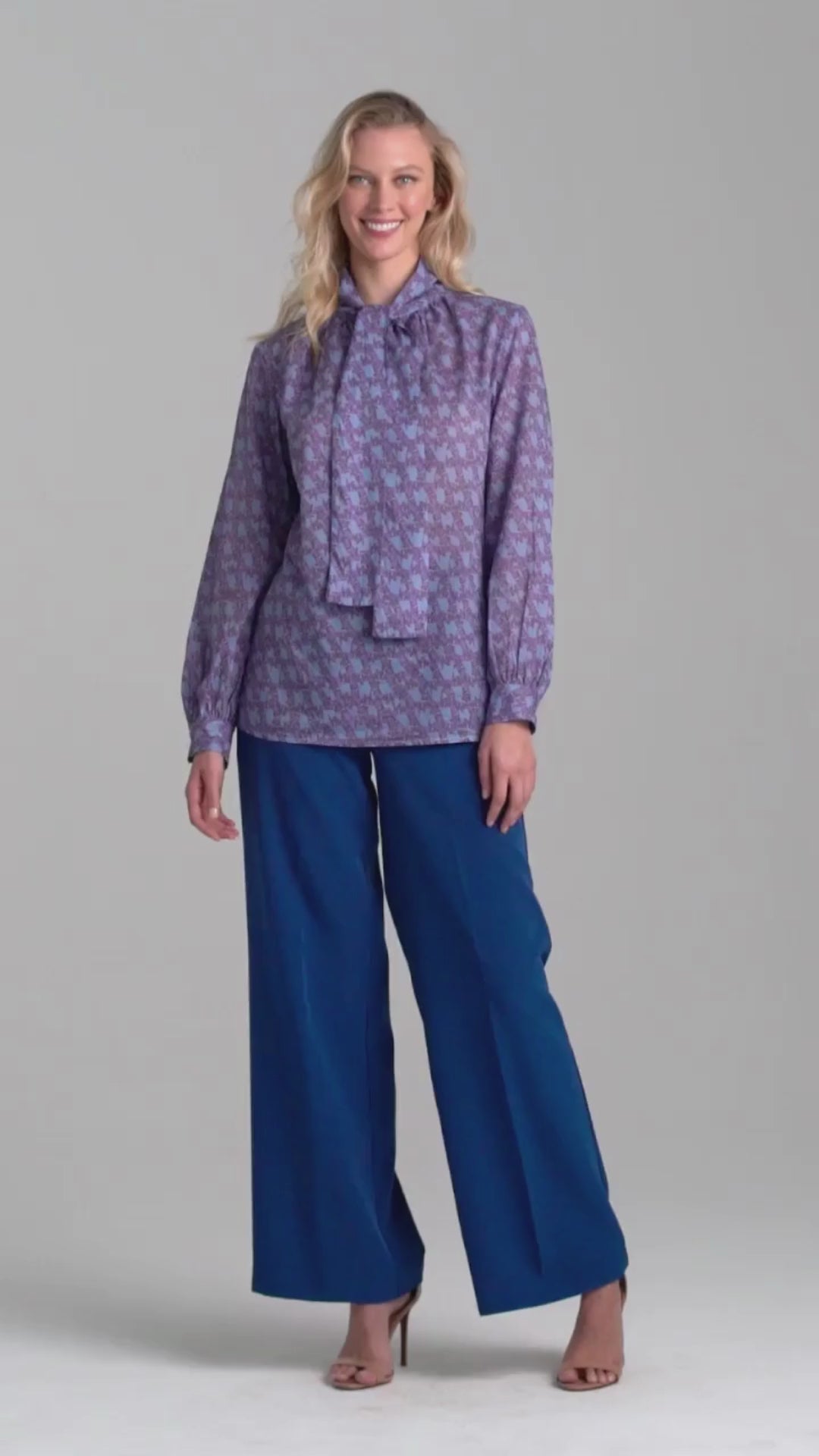 Woman wearing purple and blue bird printed cotton bow blouse with blue stretch knit pants by Ala von Auersperg for fall 2022